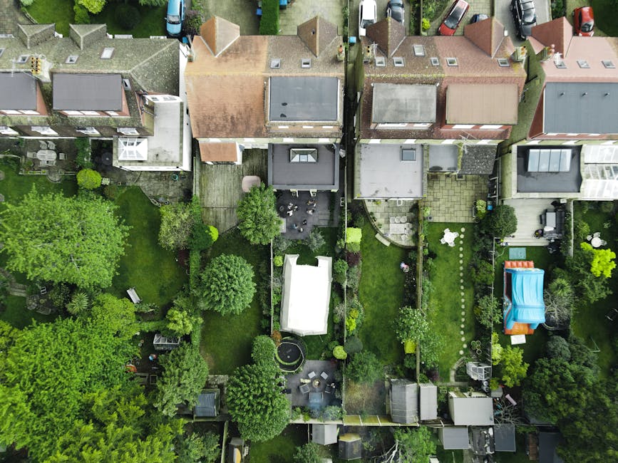 An aerial view of small city gardens viewed from above, showing various landscaped backyards behind terraced houses. The image displays a mix of green lawns, flower beds, and paved patios. Several gar