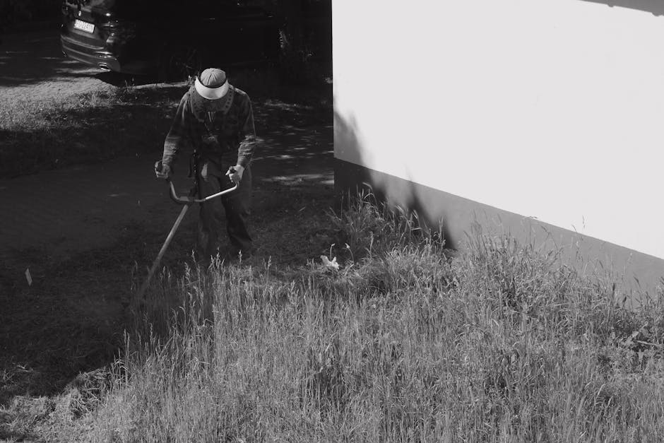 A person wearing a protective cap and jacket is using a garden hoe to cultivate or remove weeds from a patch of grassy and weedy soil near a white building wall. The garden bed features tall, untrimme