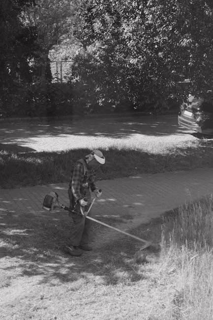 A person wearing a protective cap and jacket is using a garden hoe to cultivate or remove weeds from a patch of grassy and weedy soil near a white building wall. The garden bed features tall, untrimme